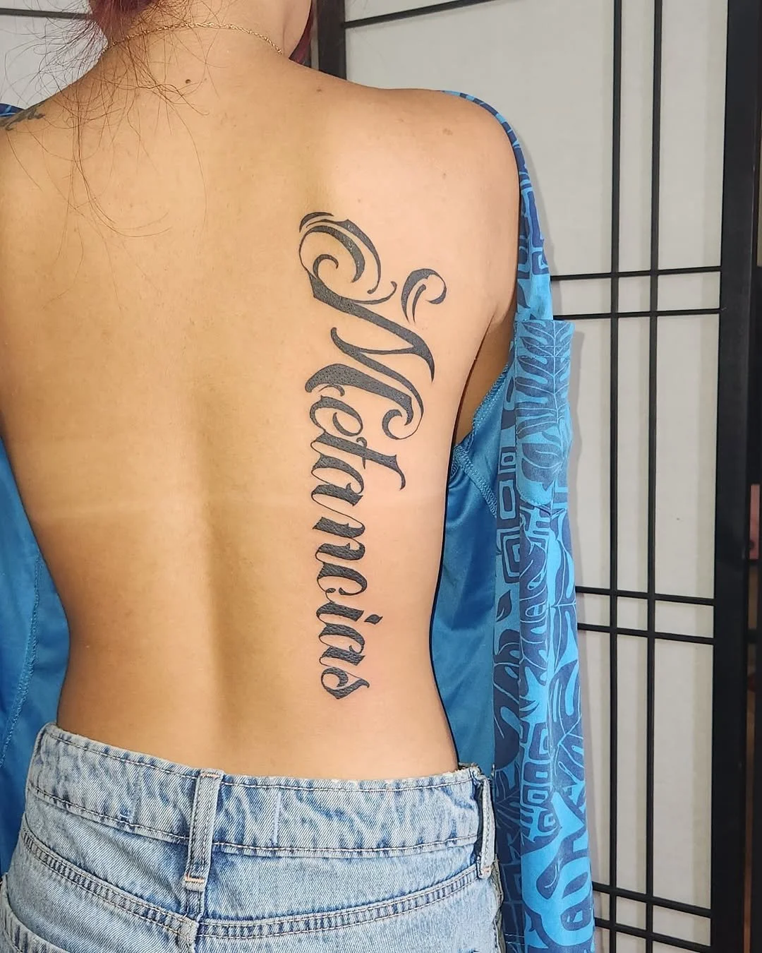 A woman with a large tattoo of the word 'Serenity' written vertically in elaborate script on her right side ribs, partially covered by a blue patterned jacket and wearing high-waisted denim shorts.