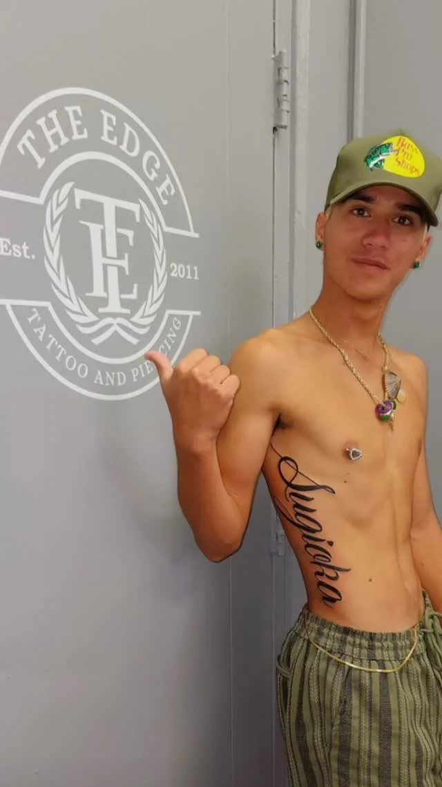 Young man with tattoos, jewelry, and a hat, shirtless, standing indoors against a wall with a logo that says 'The Edge Tattoo and Piercing,' showing a thumbs-up gesture.