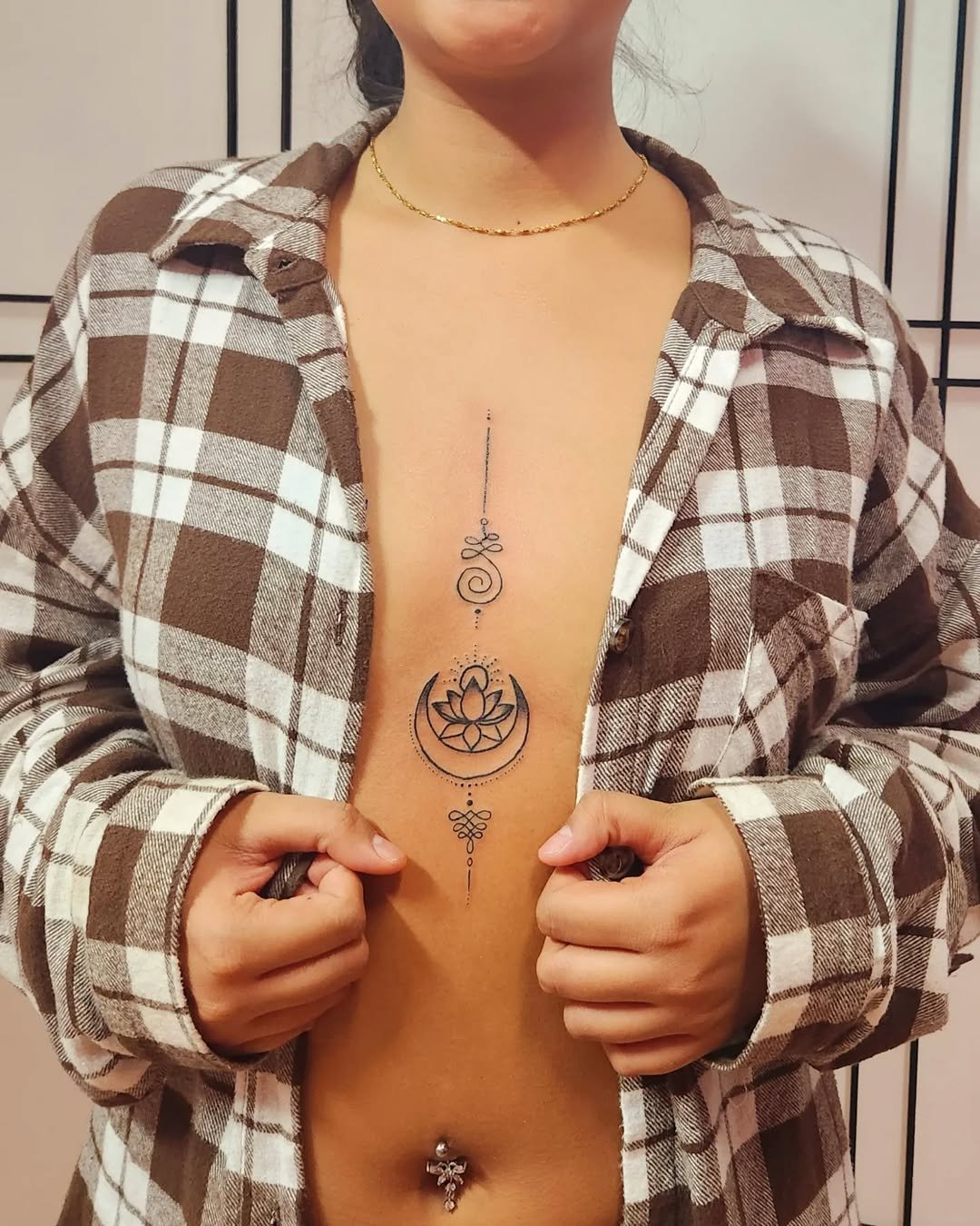 Woman in a plaid shirt showing a large vertical tattoo on her chest featuring a lotus flower inside a crescent moon with decorative elements.