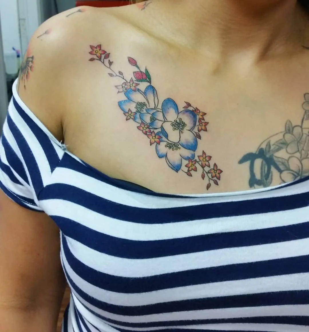 Colorful floral tattoo on person's shoulder and chest, featuring blue, pink, and yellow flowers with green leaves, and part of another tattoo visible on the side.