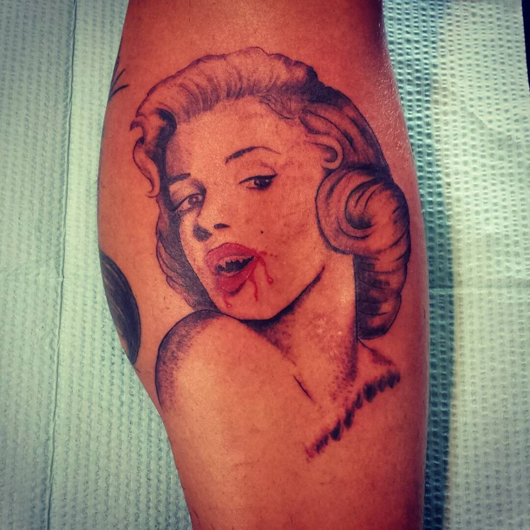 Tattoo of a woman with vintage hairstyle and red lipstick with blood on her lips, signed with the name 'Marilyn'.