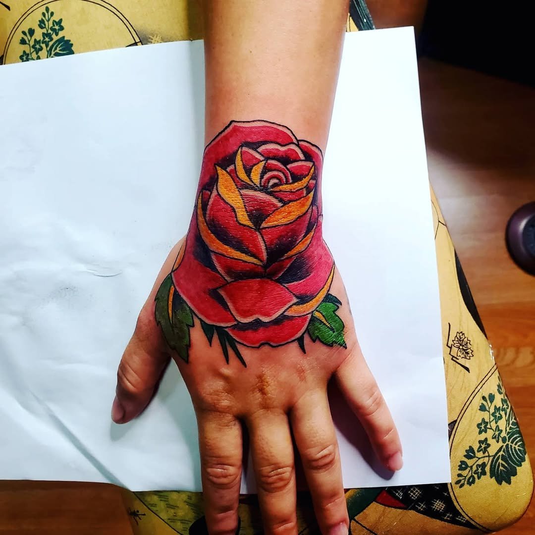 A colorful tattoo of a red rose with yellow and black accents on the hand and wrist.