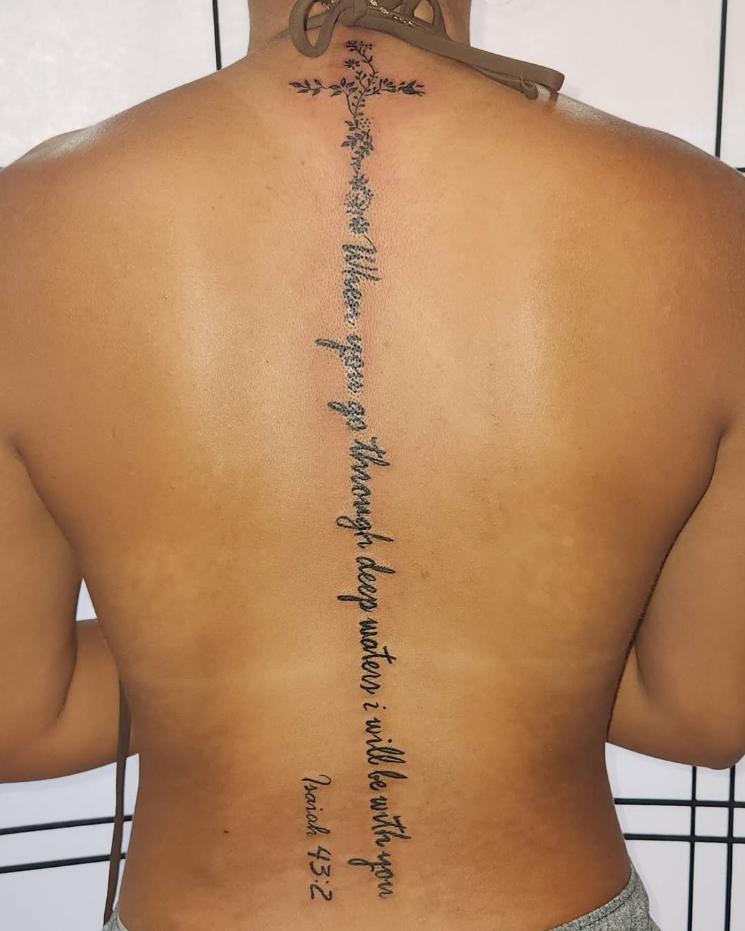 Back of a person with a vertical tattoo featuring a quote in cursive font, starting from the neck to the lower back; the person is shirtless, with a tan skin tone, and has a partial view of the face and arms.