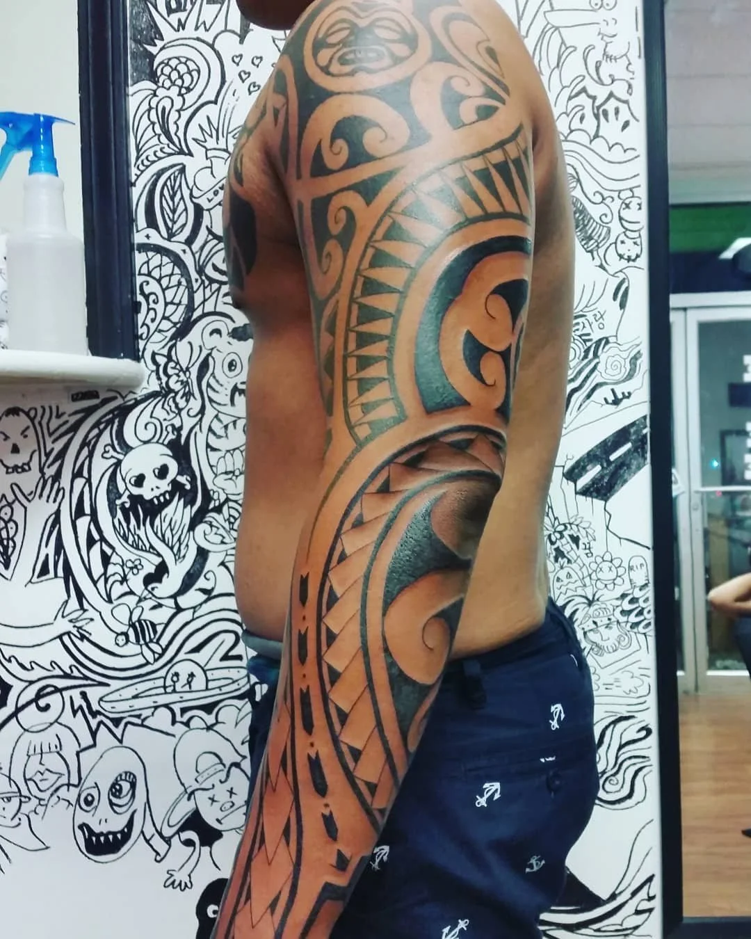 A person with a full arm Polynesian-style tattoo, featuring bold black geometric tribal patterns extending from shoulder to wrist.