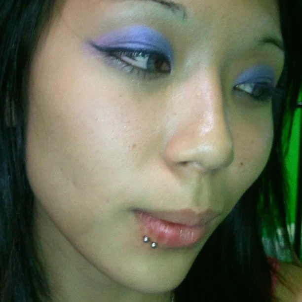 Close-up of a young woman with purple eyeshadow, dyed eyebrows, and a lip piercing.