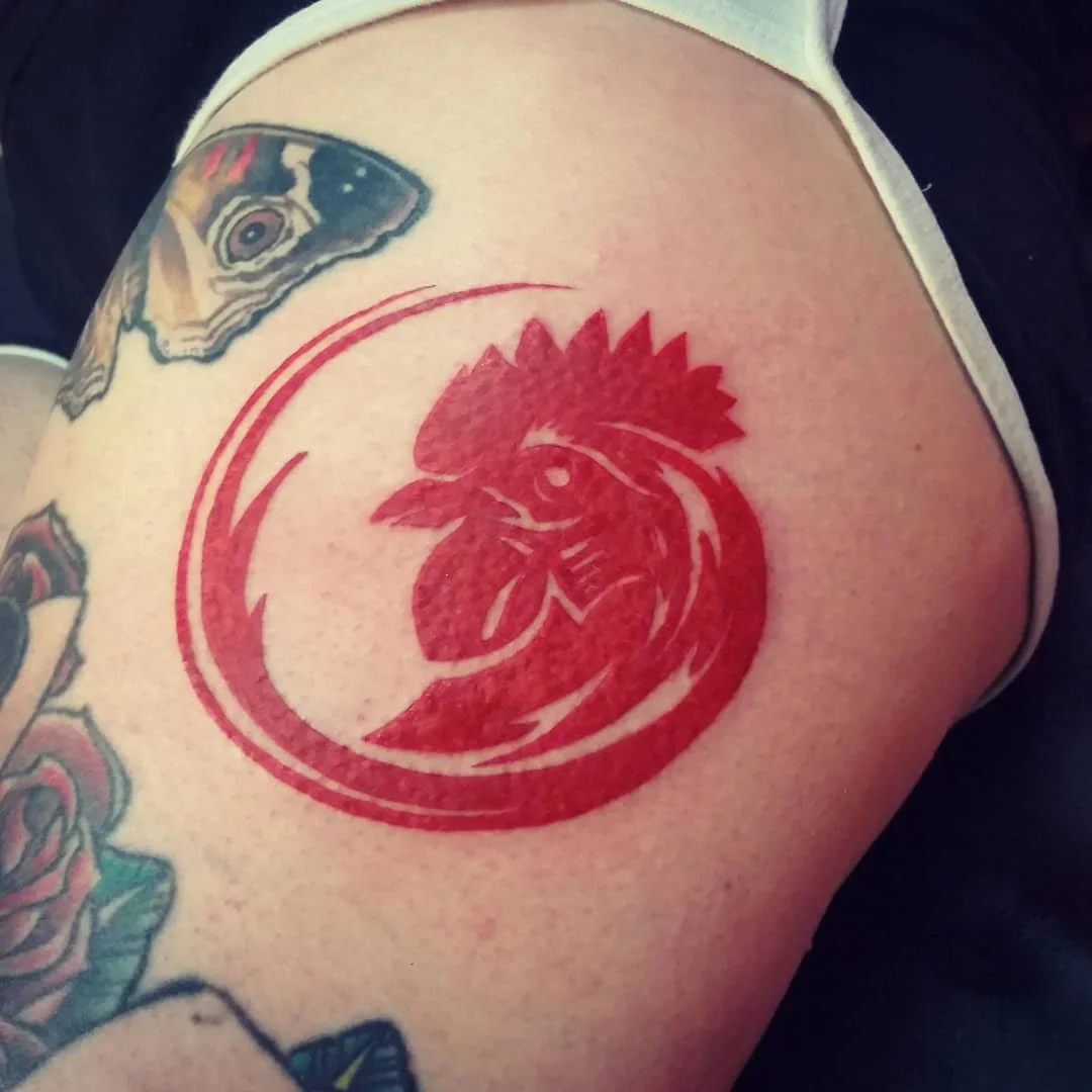 Red tattoo of a rooster head inside a circular border on a person's upper arm, surrounded by other tattoos, including a butterfly and roses.