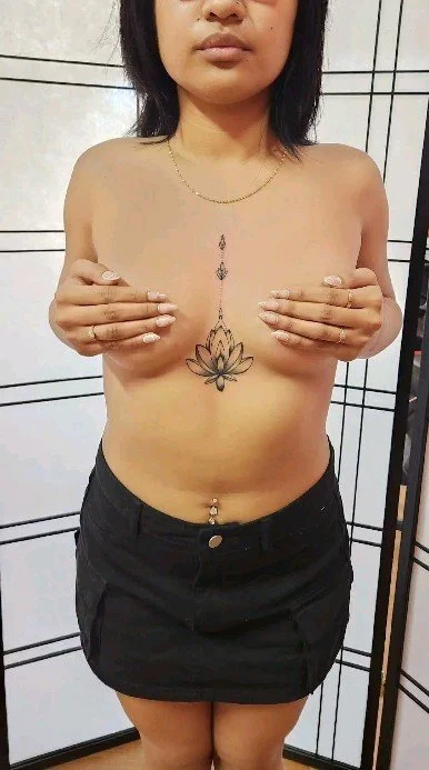 Woman with a lotus flower tattoo on her chest covering her breasts, standing in front of a tiled wall.