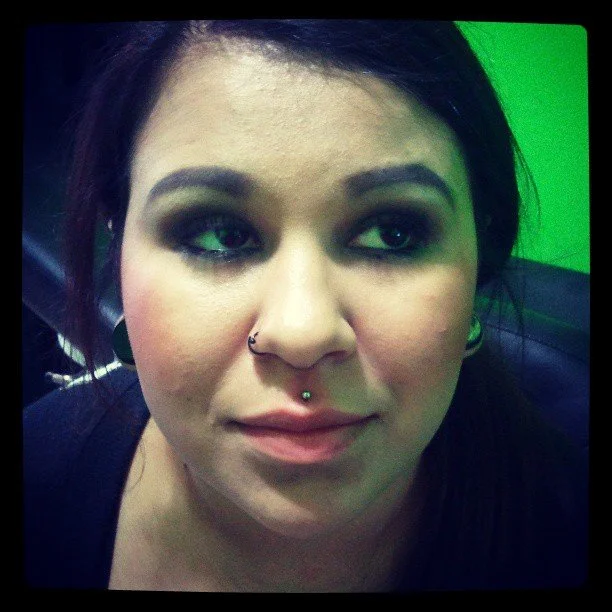 Close-up of a woman with dark hair, multiple facial piercings, makeup, and a slight smile, against a green background.