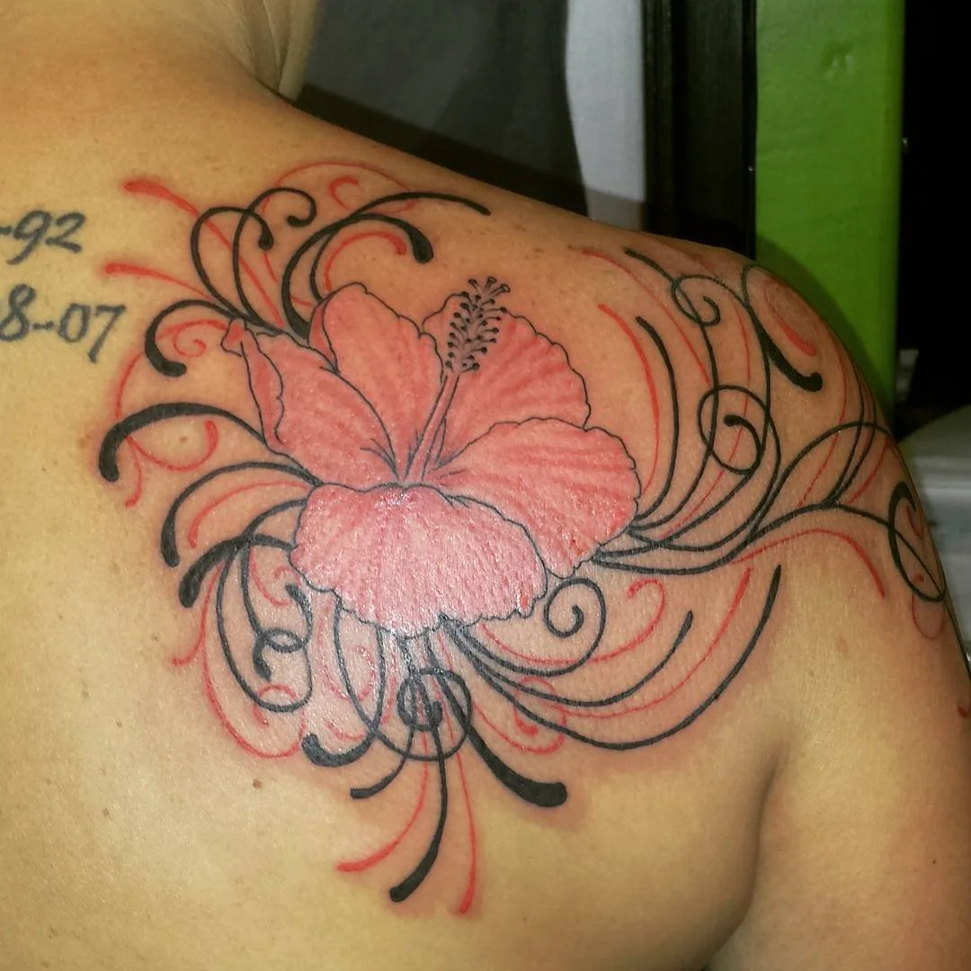 A tattoo of a pink hibiscus flower with black and red decorative swirls on a person's shoulder.