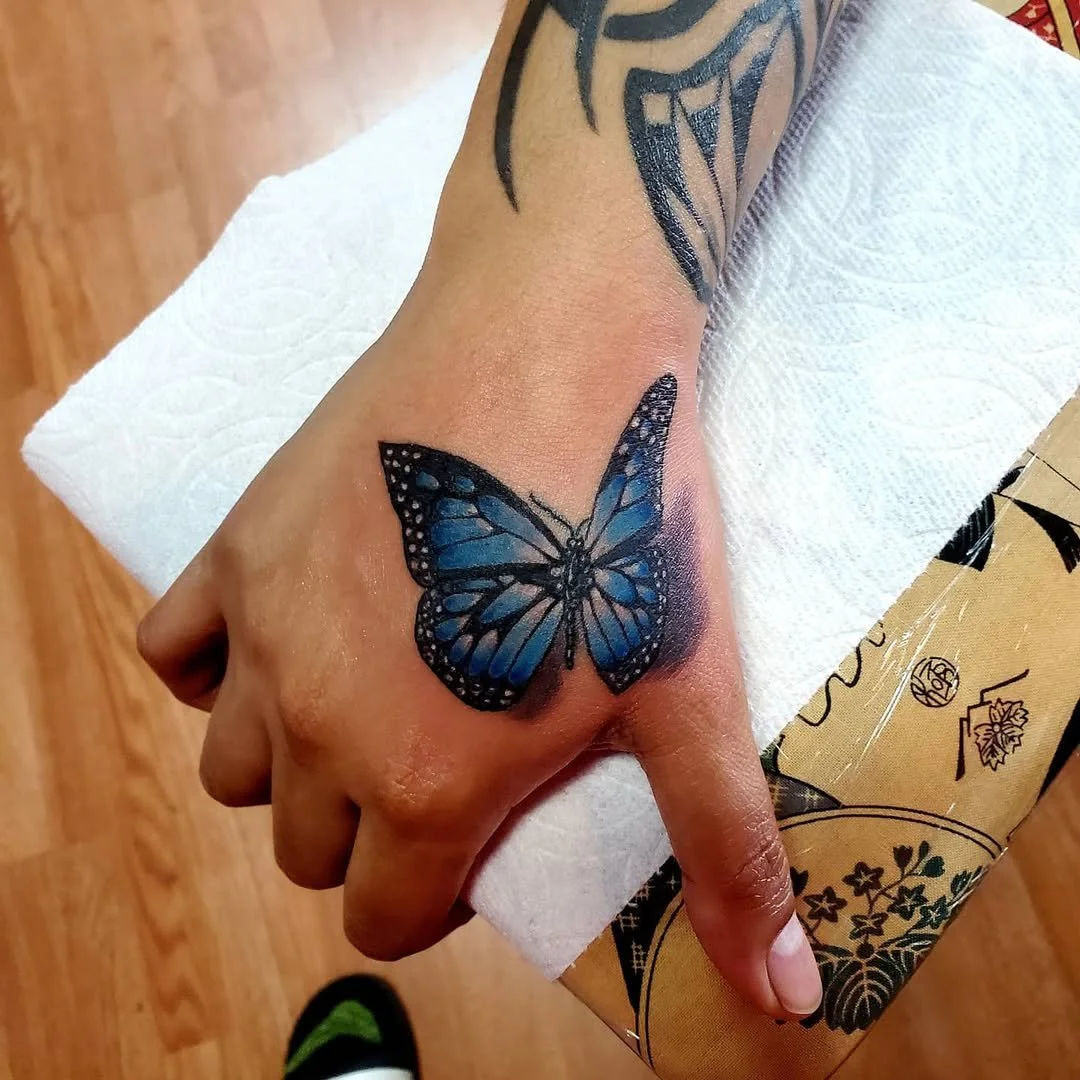 A freshly inked blue butterfly tattoo on a person's hand with detailed black outline and shading, resting on a white paper towel.