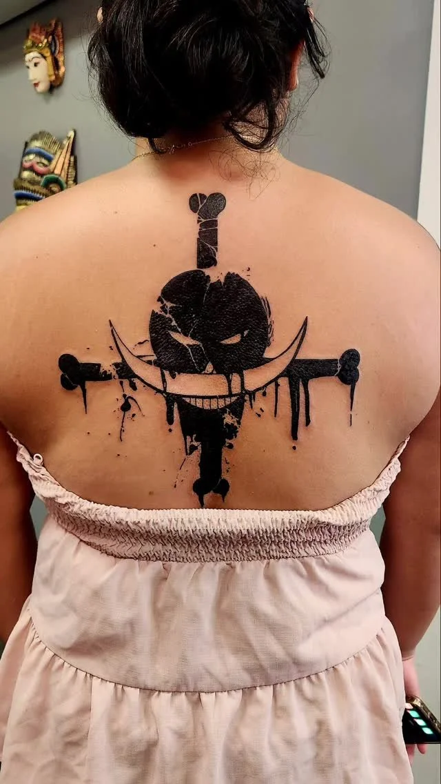 A woman with black hair in a towel showcases a black ink tattoo on her upper back, depicting a skull and crossbones with a sword through the skull, rendered in a splattered, graffiti style.