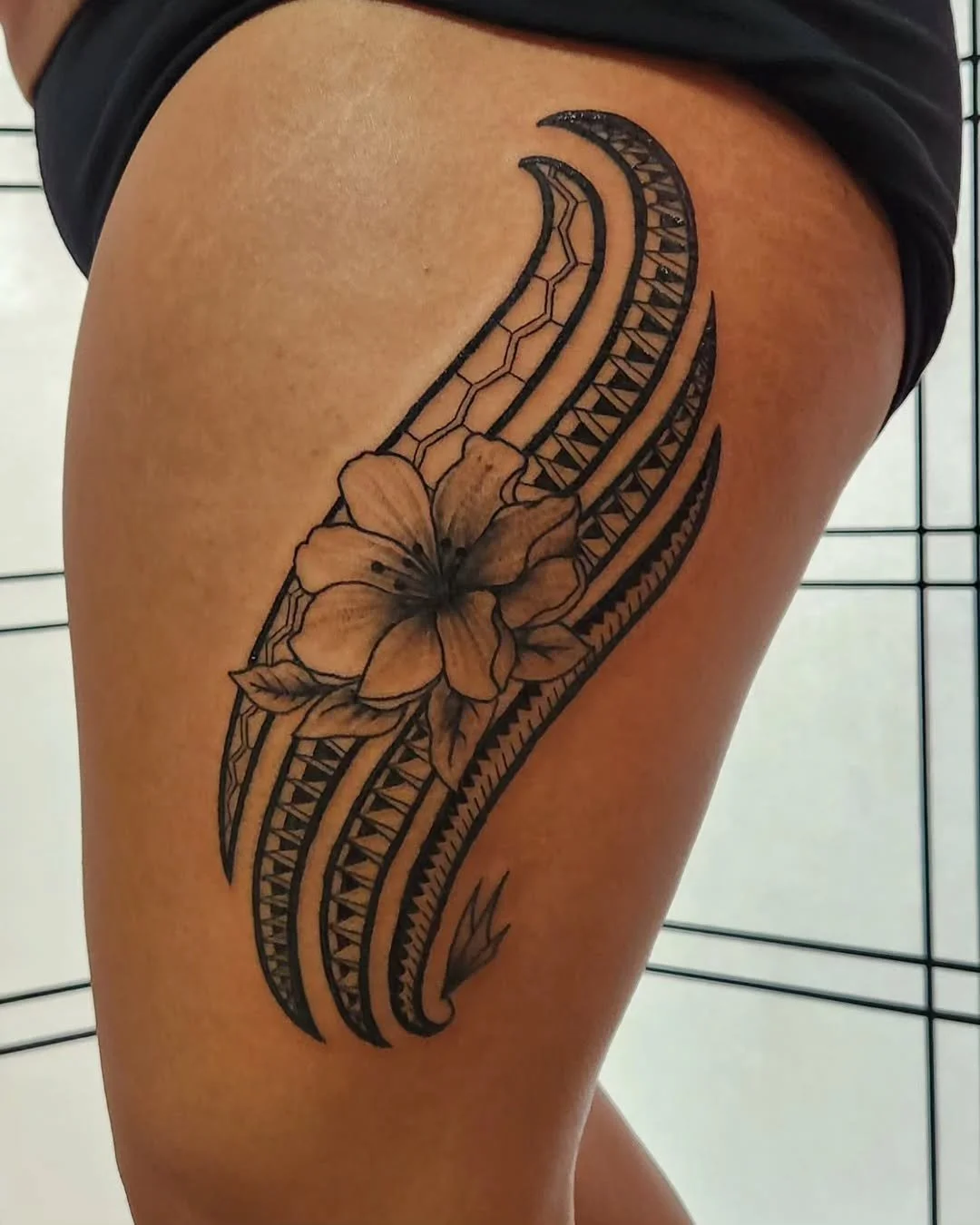 A black ink tattoo on a person's thigh featuring tribal patterns intertwined with a detailed hibiscus flower.