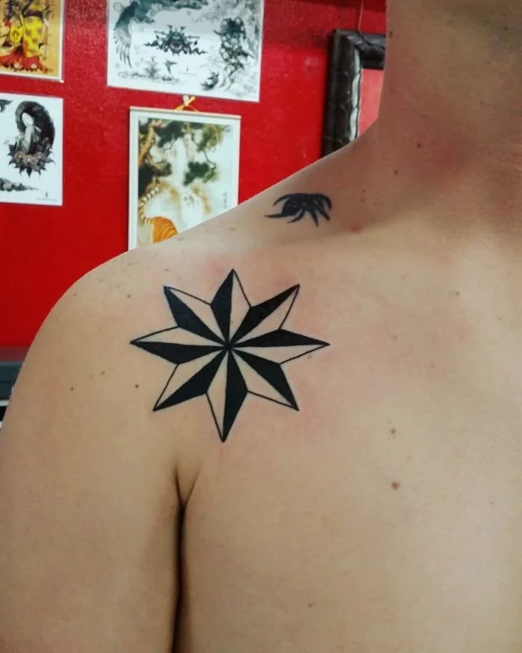 A person has a black star-shaped tattoo on their shoulder with sixteen points and bold black fill. There is a smaller tattoo of a black creature on the neck, possibly a lizard or salamander. The background features a red wall with various art prints 