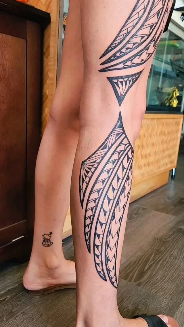 A person's leg with a large, intricate black Polynesian-style tribal tattoo extending from the thigh to the ankle. There is also a small tattoo of a teddy bear near the ankle.