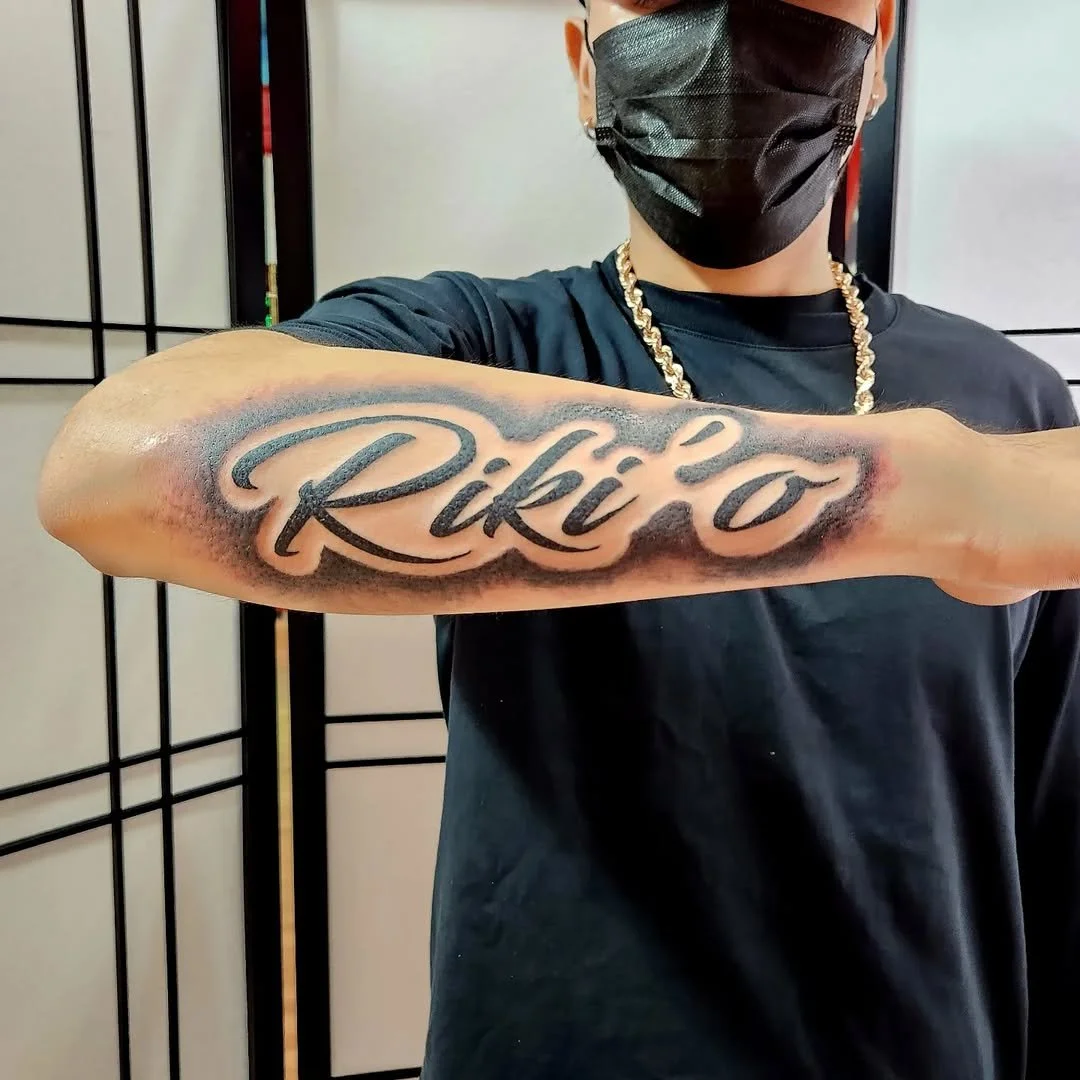 A person with a face mask, earrings, and a gold chain, displaying a tattoo on their forearm that says 'Rikko' in cursive script.