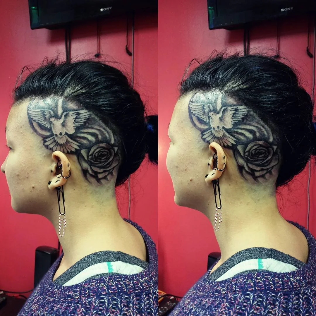 Side view of a person with dark hair styled in a bun, featuring a large tattoo of a dove and a rose on the side of their head, with multiple earrings and a purple patterned sweater.