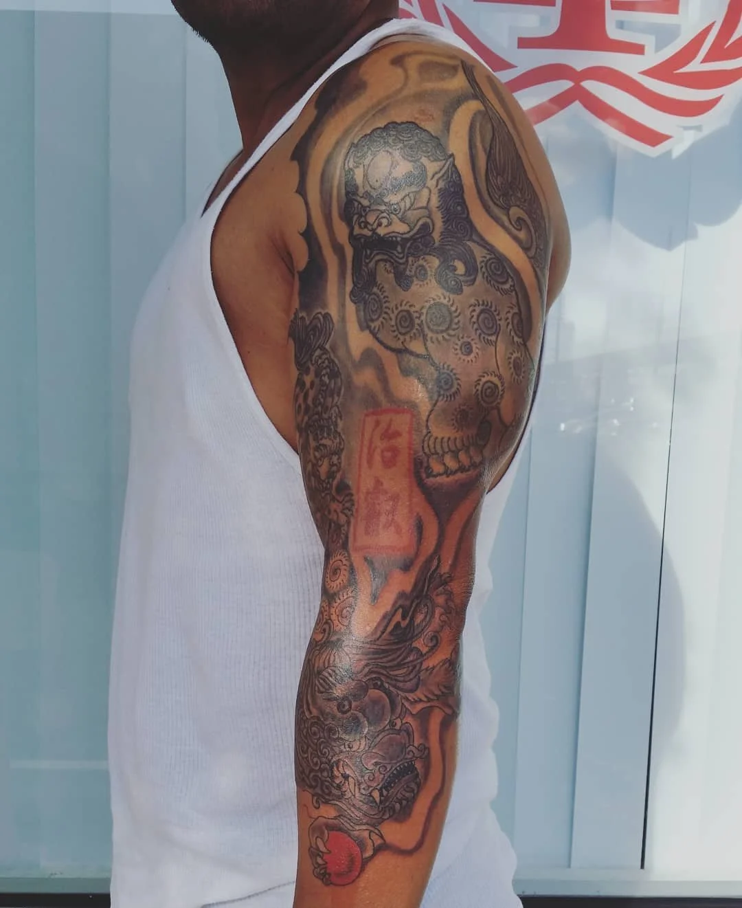 Tattooed man's arm with a dragon, a skull, and red and black ink on a white sleeveless shirt.