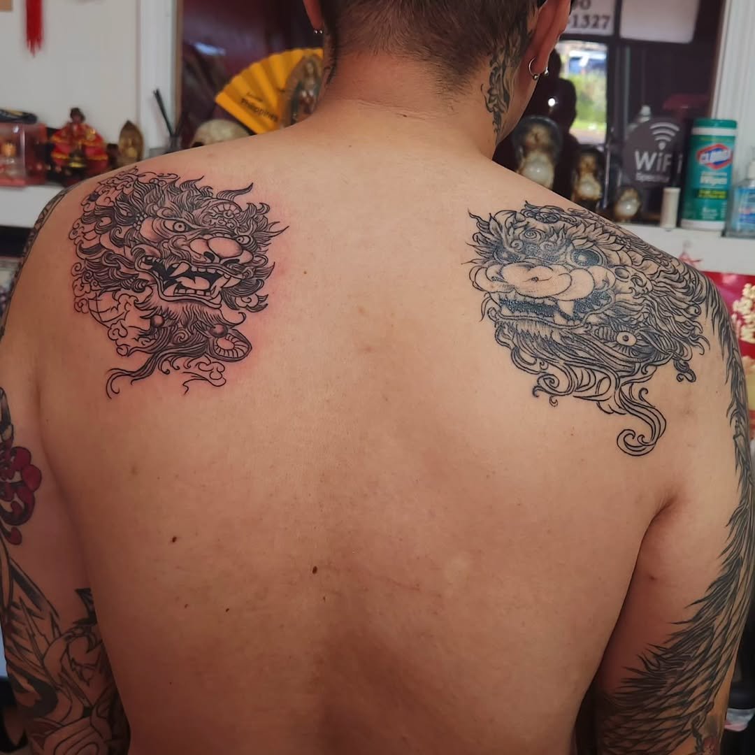 Tattooed man with two Asian-style dragon face tattoos on shoulders, tattoo shop background.