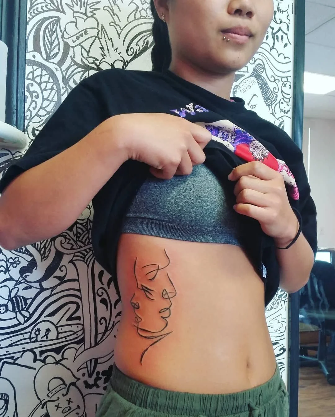 A woman is lifting her black T-shirt to reveal a tattoo on her side, depicting a line art profile of a woman's face. She is standing in front of a black and white mural with various drawings.