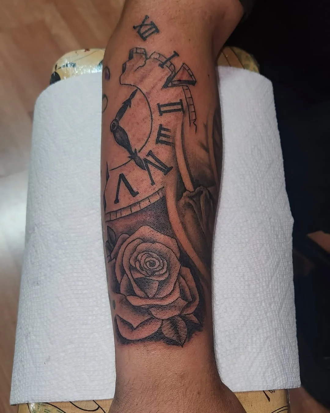A black and gray tattoo on a person's forearm featuring a clock with Roman numerals, a rose, and decorative elements.