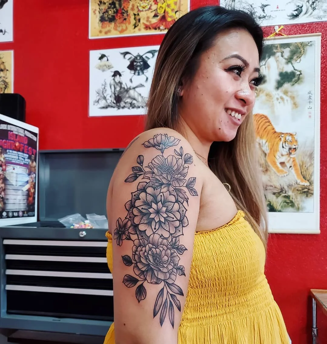 A woman with long hair wearing a yellow strapless dress showing a floral tattoo on her upper arm, smiling and looking to the right, with colorful Asian-style art and a tiger painting in the background.