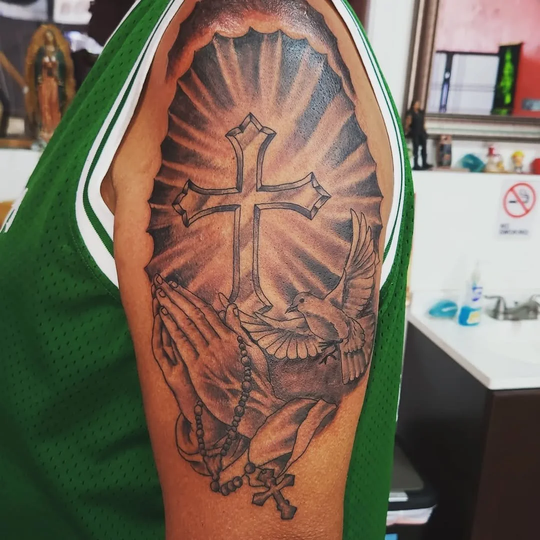 Tattoo of praying hands, rosary beads, a dove, a cross, and a glowing cross in the background on a person's upper arm.