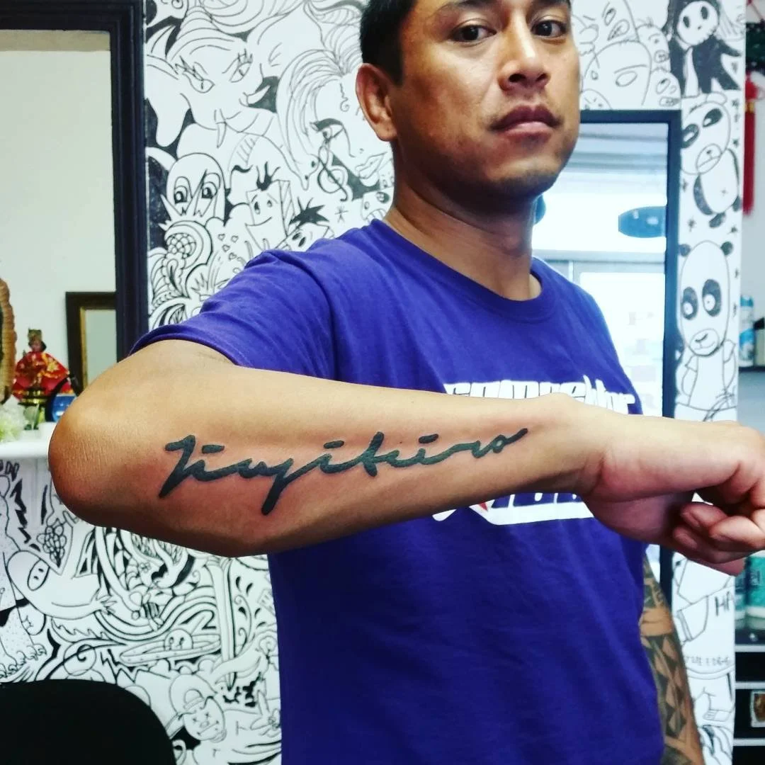 A man with short dark hair wearing a purple T-shirt showing a tattoo that says 'Tokyo' on his forearm, standing in a tattoo studio with black and white cartoon-style wall art in the background.