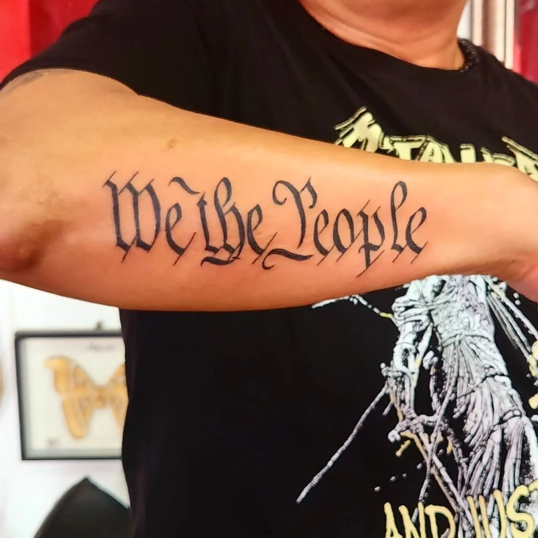 Tattoo says 'We the People' on a person's upper arm.