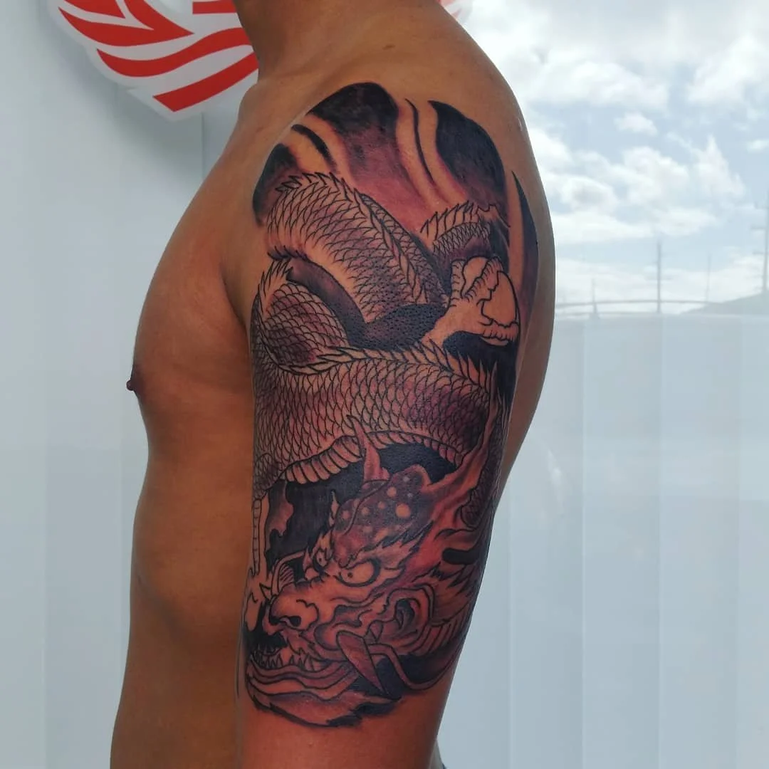 A tattoo of a dragon on a person's upper arm and shoulder, with a background of clouds and sky.