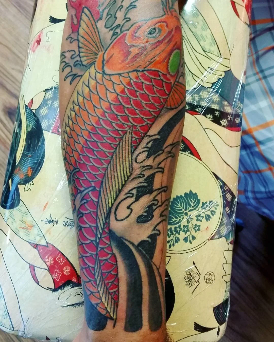 A colorful tattoo of a koi fish with orange and red scales, green accents, and detailed black outlines on a person's forearm.