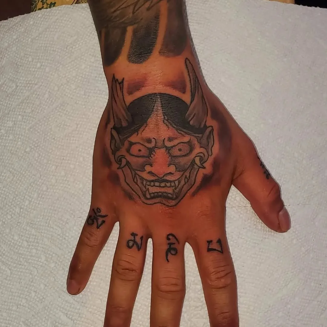 Tattooed hand with a large demon face tattoo and numbers on the fingers.