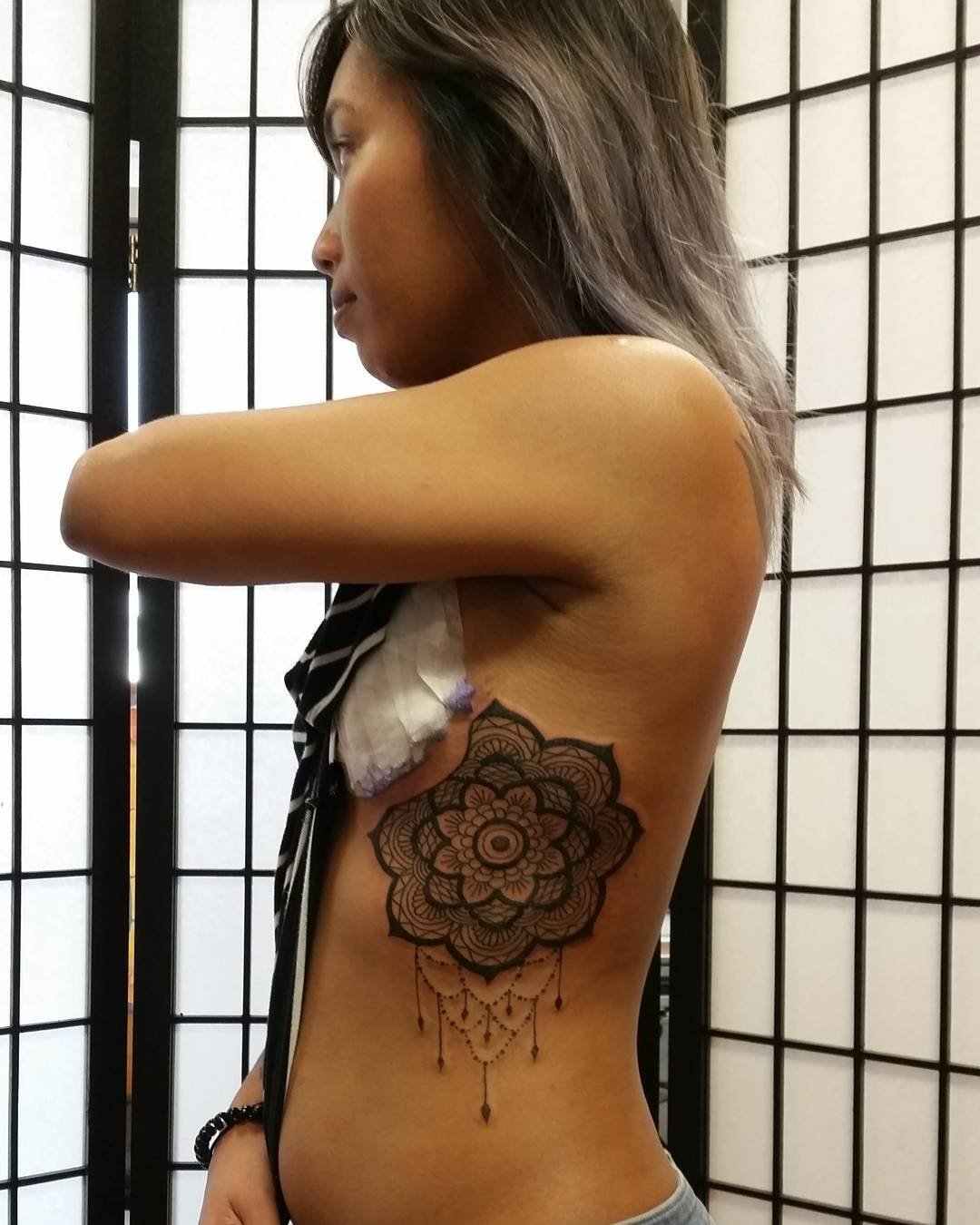 A woman with brown hair and a tattoo of a floral mandala with hanging chains on her side, standing in front of a black and white paper screen.