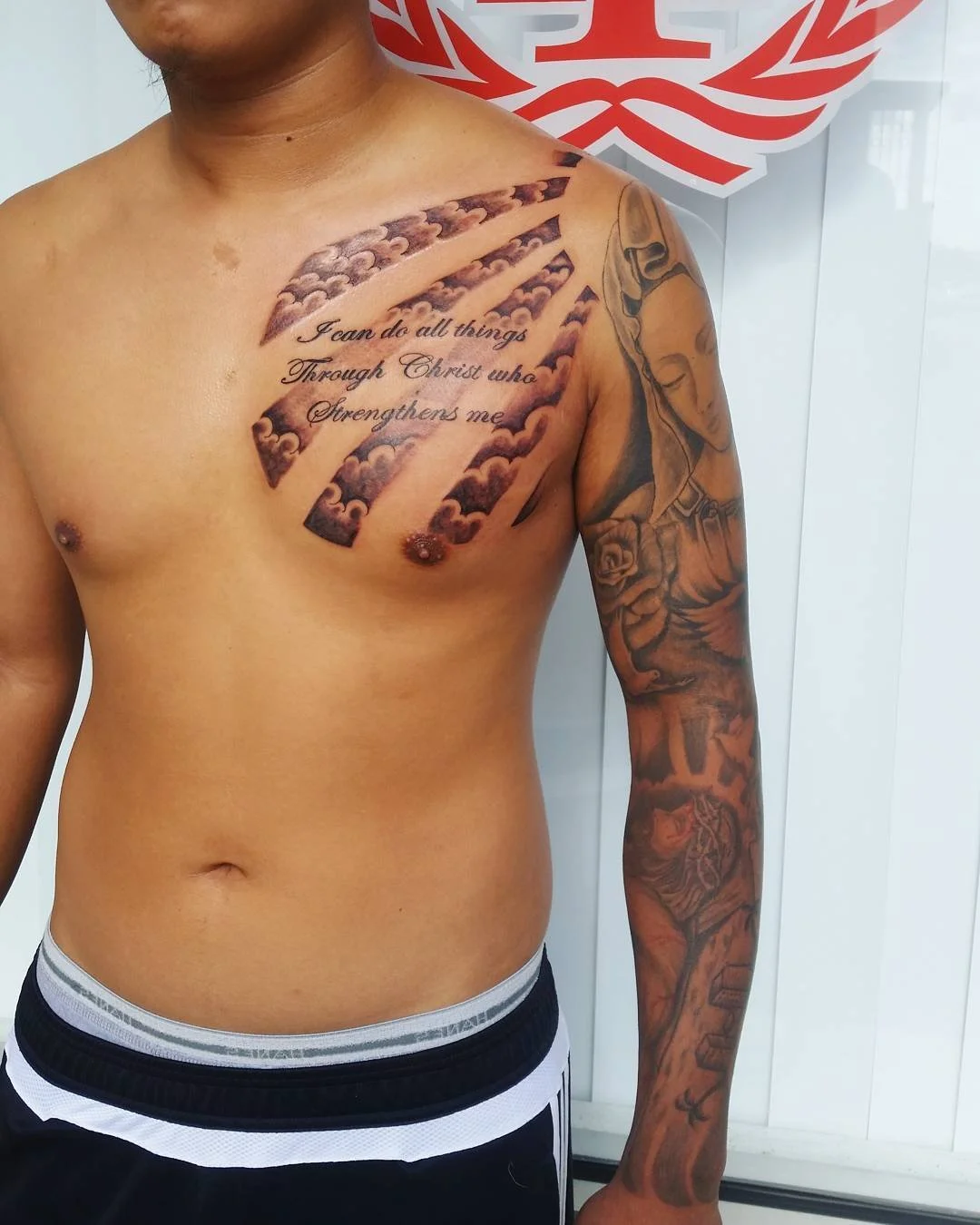 A shirtless man with tattoos on his chest and right arm, standing in front of a white wall with a red and white emblem.