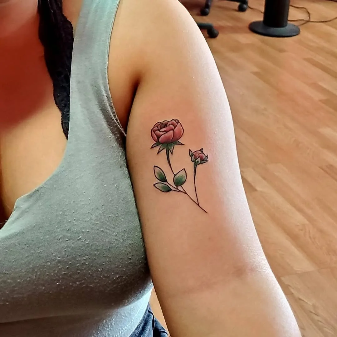 A close-up of a person's upper arm tattoo depicting a pink and red rose with green leaves. The person is wearing a sleeveless shirt and is indoors with wooden flooring and some chairs and tables in the background.