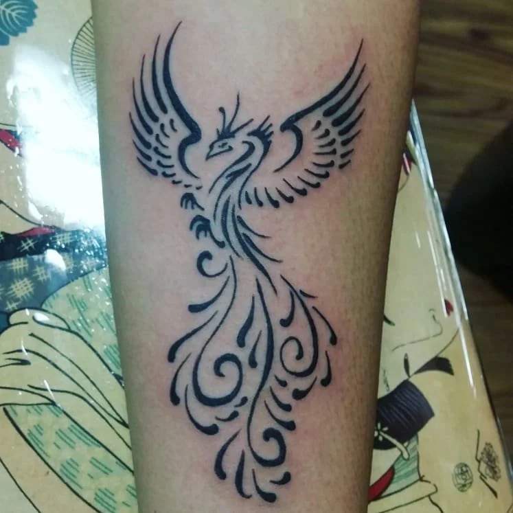 Tattoo of a stylized eagle with outspread wings and elongated tail feathers on a person's arm.