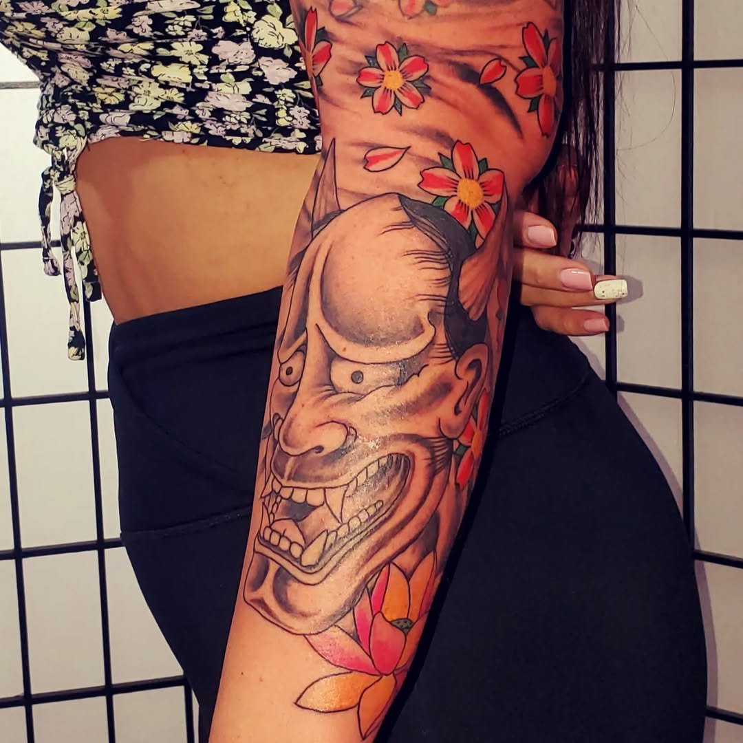 Tattoo of a traditional demon or oni mask with sharp teeth, expressive eyebrows, and a prominent nose, surrounded by colorful flowers on the person's arm.