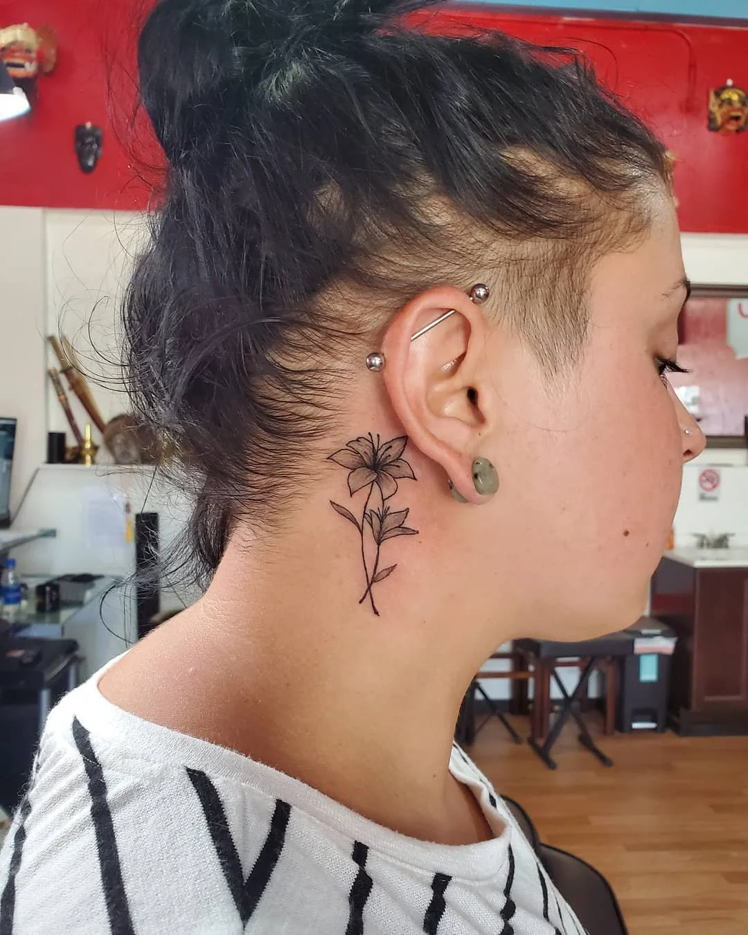 Side view of a woman with dark hair, an undercut hairstyle, multiple piercings, and a floral tattoo behind her ear. She is wearing a white shirt with black stripes, and the background shows a room with red walls and various decor.