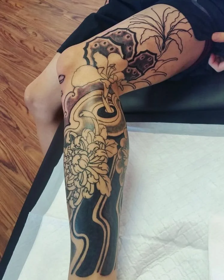A person getting a tattoo on their thigh, featuring floral and leaf designs with some shaded sections.