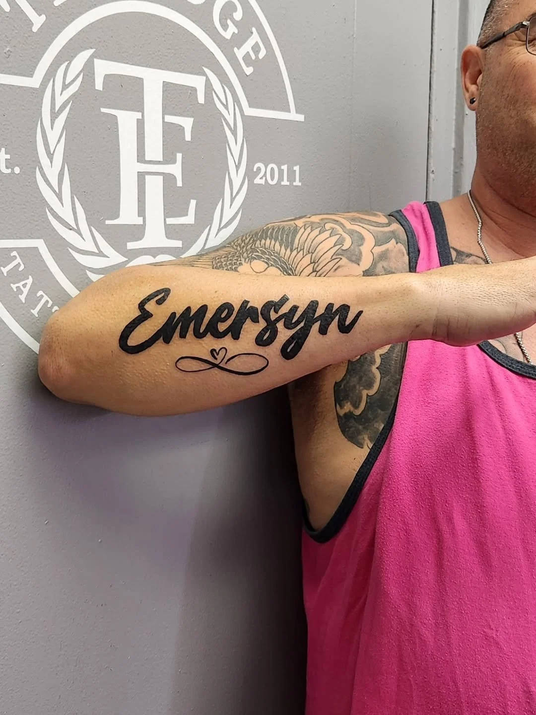 A person with tattoos on their arm, wearing a pink sleeveless shirt, displays a tattoo with the word 'Emersyn' in black cursive font on their inner arm.