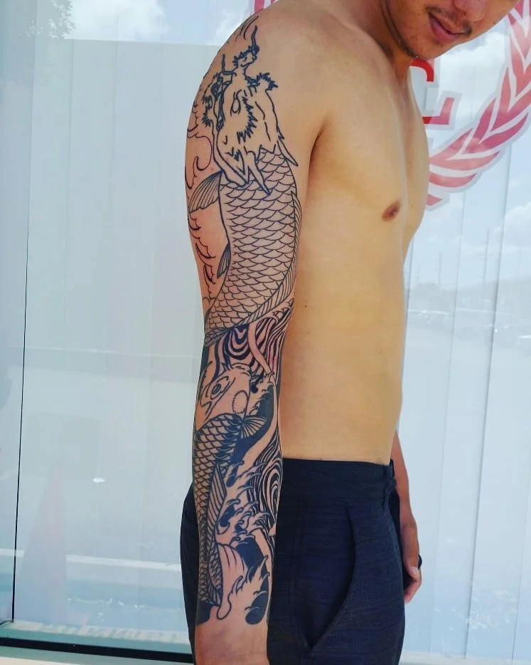 A shirtless man with a large, detailed tattoo of a dragon and waves on his left arm, standing indoors near a window with a slight smile.