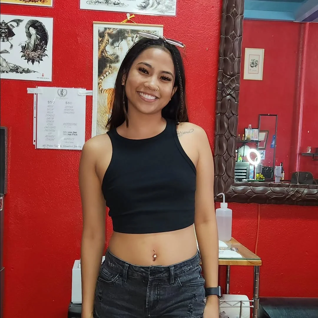 A young woman with long dark hair, wearing a black crop top and dark jeans, smiling and standing in a colorful tattoo shop with a red wall and various framed artwork.