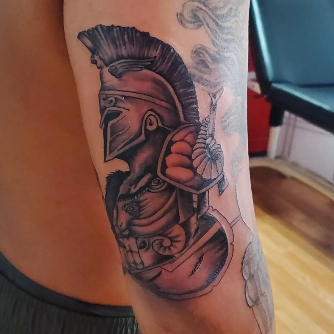 Tattoo of a Greek or Roman soldier wearing a helmet with a crest, holding a shield, with decorative elements and smoke, in black and gray ink.