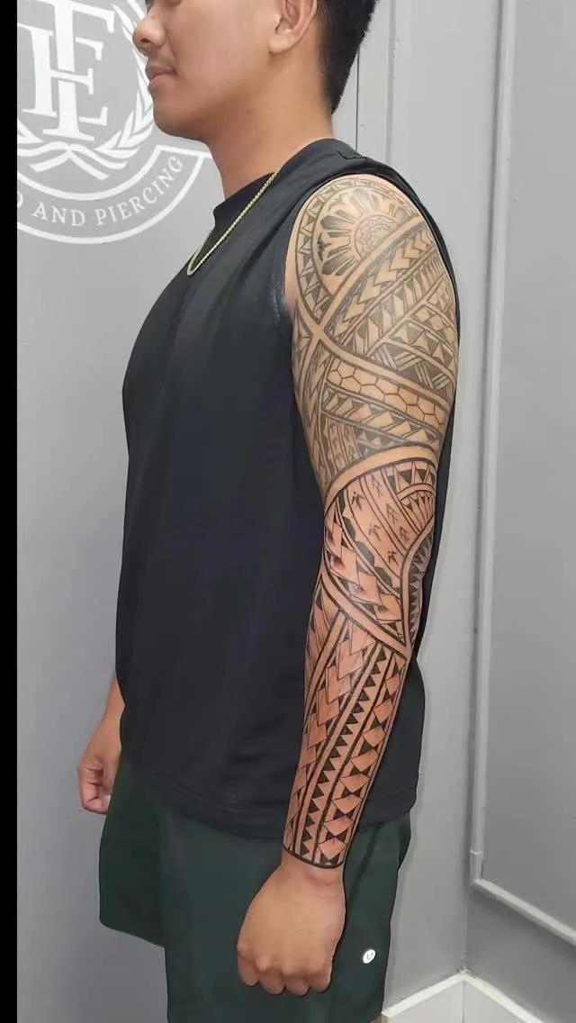 A young man with a Polynesian tattoo sleeve on his left arm, wearing a black sleeveless shirt, standing in front of a gray wall with a logo.