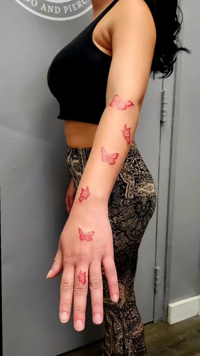 A woman with butterfly tattoos on her left arm, wearing a black top and patterned pants, standing in front of a gray wall.