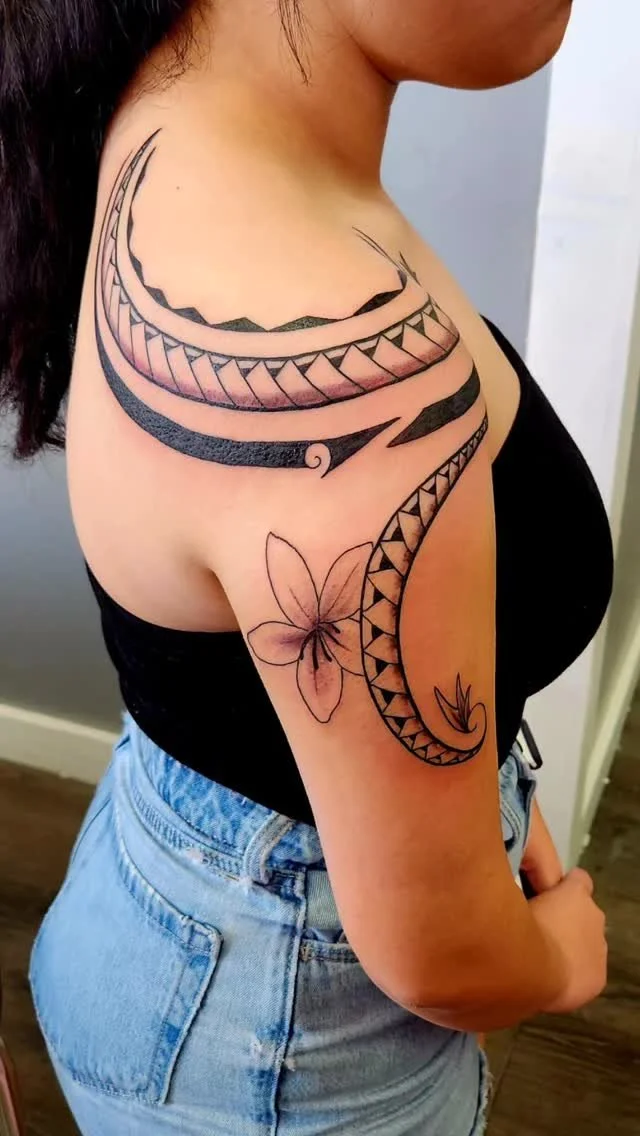 Tattoo of tribal patterns and geometric designs on the shoulder and upper arm, with a realistic flower outline.