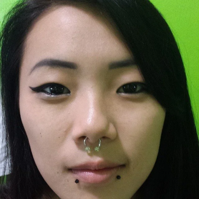 Close-up of a woman with black hair, winged eyeliner, septum piercing, and two lip piercings, against a green background.