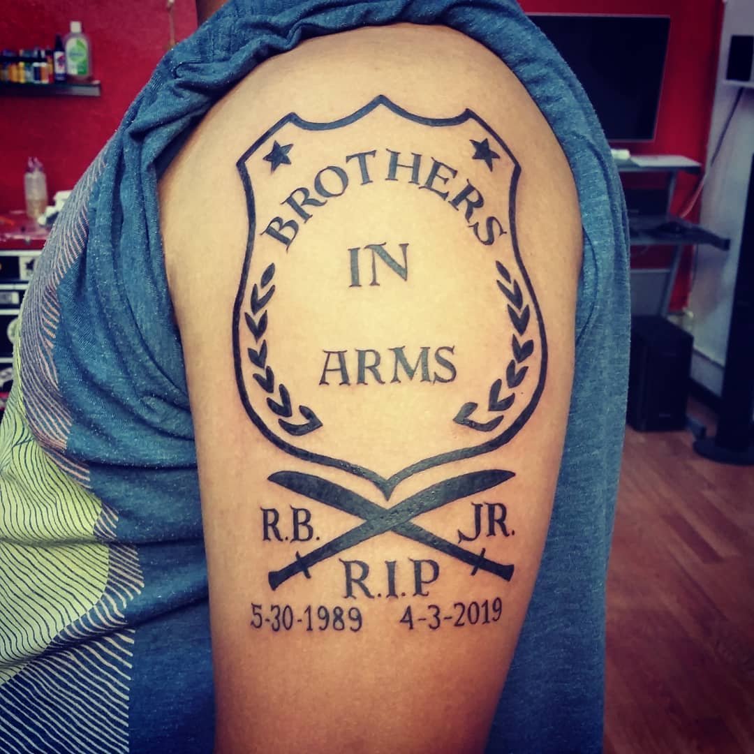 Tattoo on person's upper arm that reads 'Brothers in Arms' inside a shield shape, with two crossed swords below, along with initials RB and JR, and dates 5-30-1989 and 4-3-2019.