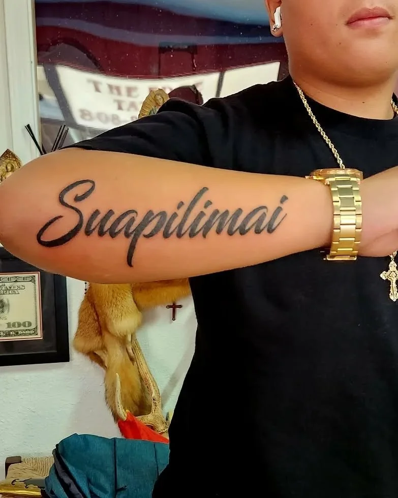 A person with a tattoo on their forearm that reads 'Suapilimai' in black cursive letters. The person is wearing a black shirt, a gold watch, and a gold chain with a cross pendant. The background includes a framed $100 bill and a decorative object res