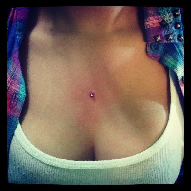 Close-up of a person's upper chest with a small heart-shaped piercing in the center, wearing a white tank top and a colorful plaid shirt.