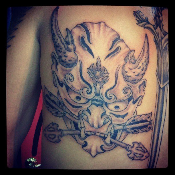Black and gray tattoo of a fierce demon or oni face with horns, a dragon in the center of its forehead, and sharp features, with a sword crossing behind the face.